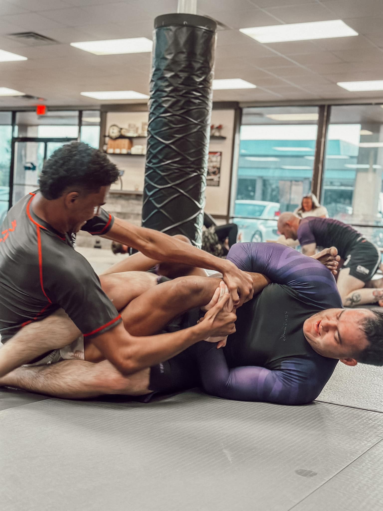 Two martial artists in a grappling match on the mat, one securing a leg lock against his opponent.