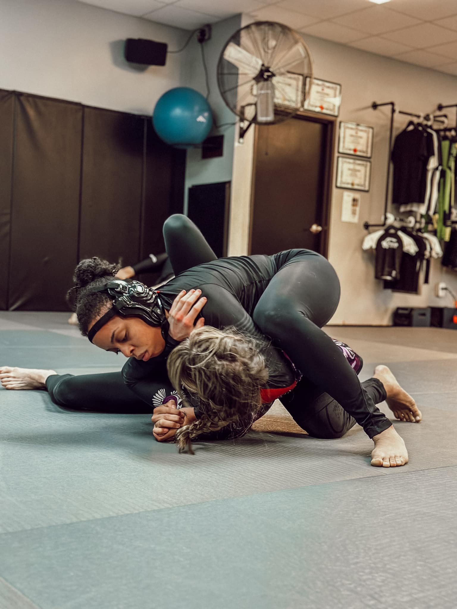 Two individuals engaged in a Brazilian Jiu-Jitsu training session on the gym floor, with one in a dominant top position.