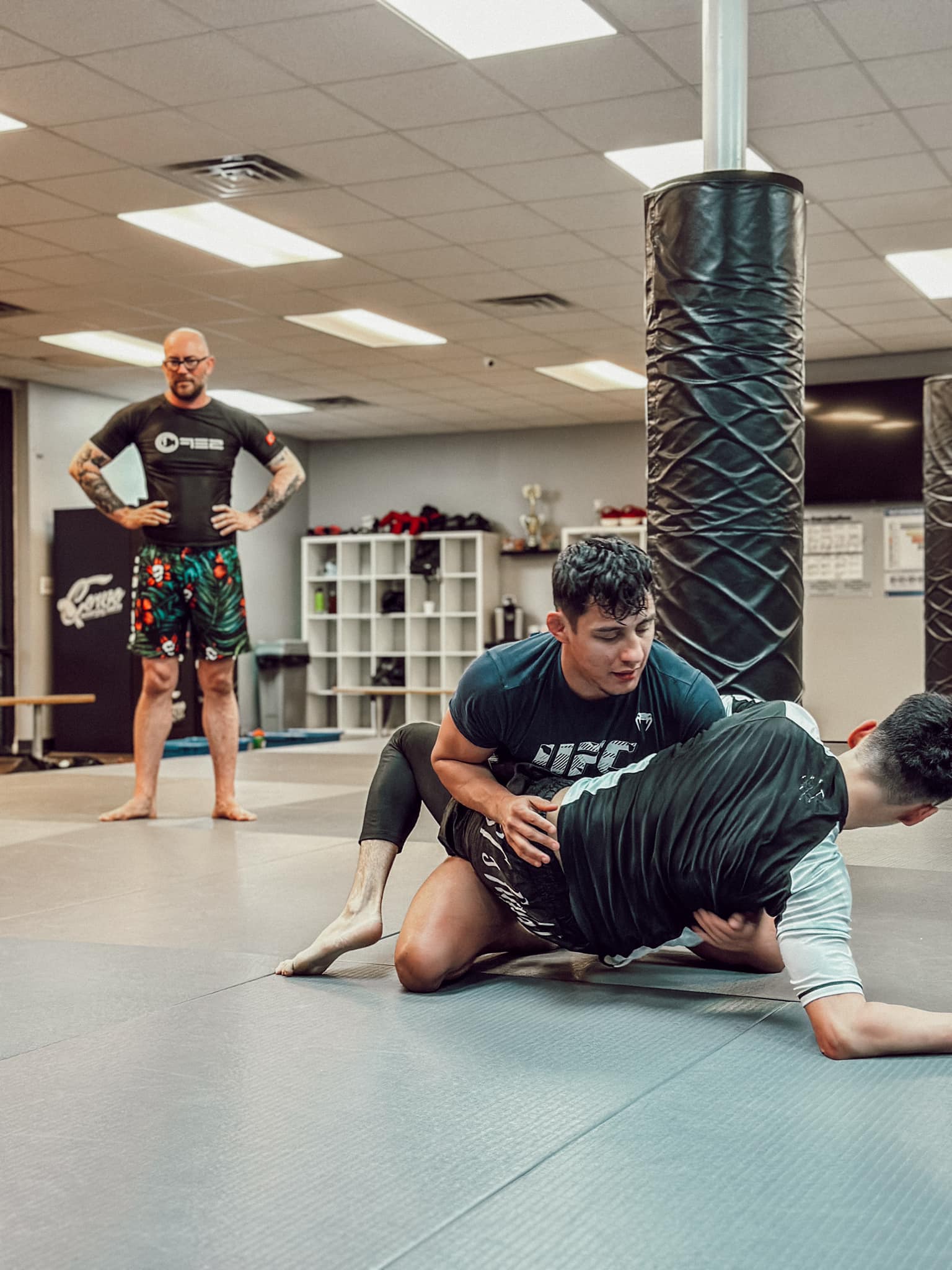 Two individuals practice Brazilian Jiu-Jitsu on the mat while a coach supervises in the background.