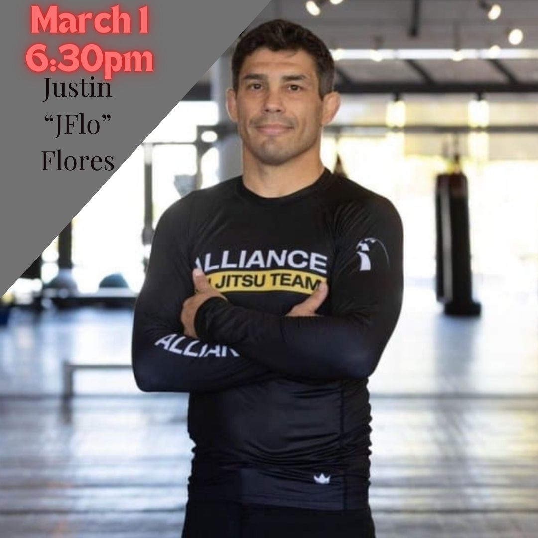 Justin 'JFlo' Flores standing with arms crossed in a martial arts gym, wearing a black Alliance Jiu-Jitsu team rashguard.